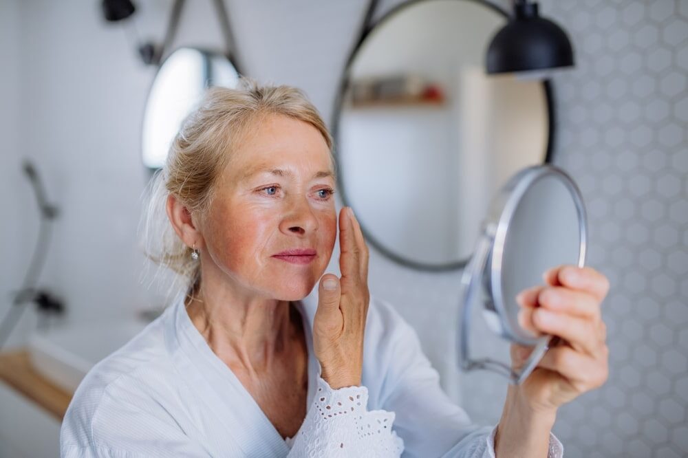 Beautiful senior woman in bathrobe looking at mirror