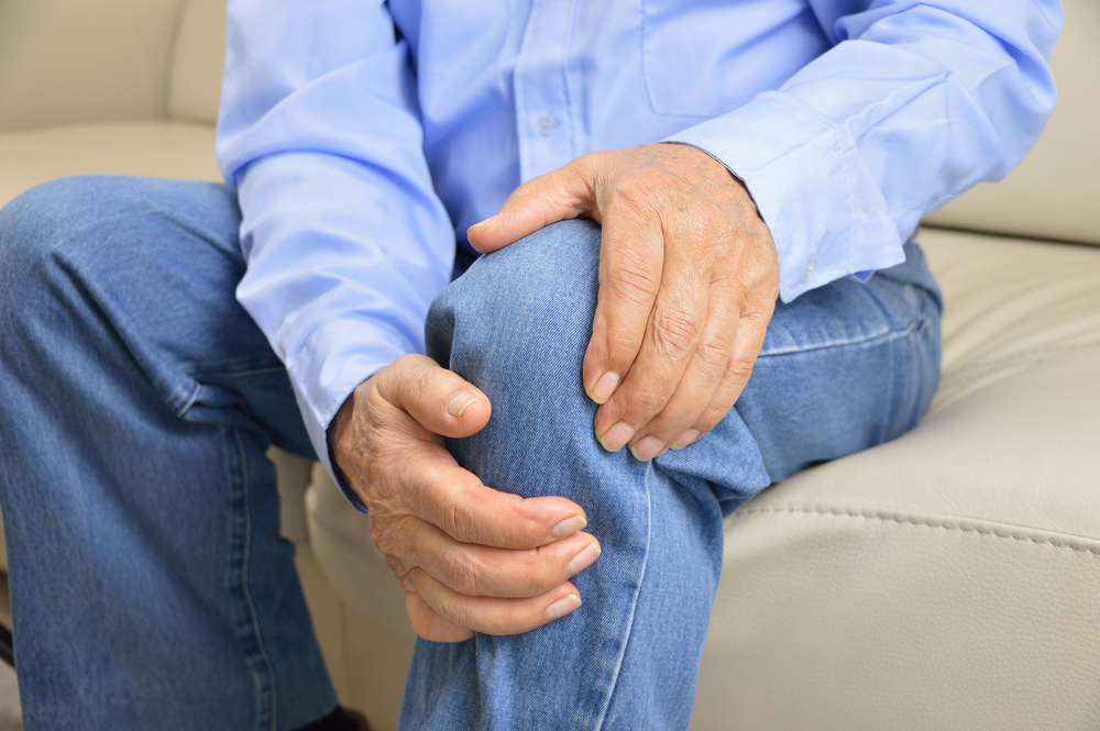 elderly man sitting on a sofa in the living room at home and touching his knee by the pain