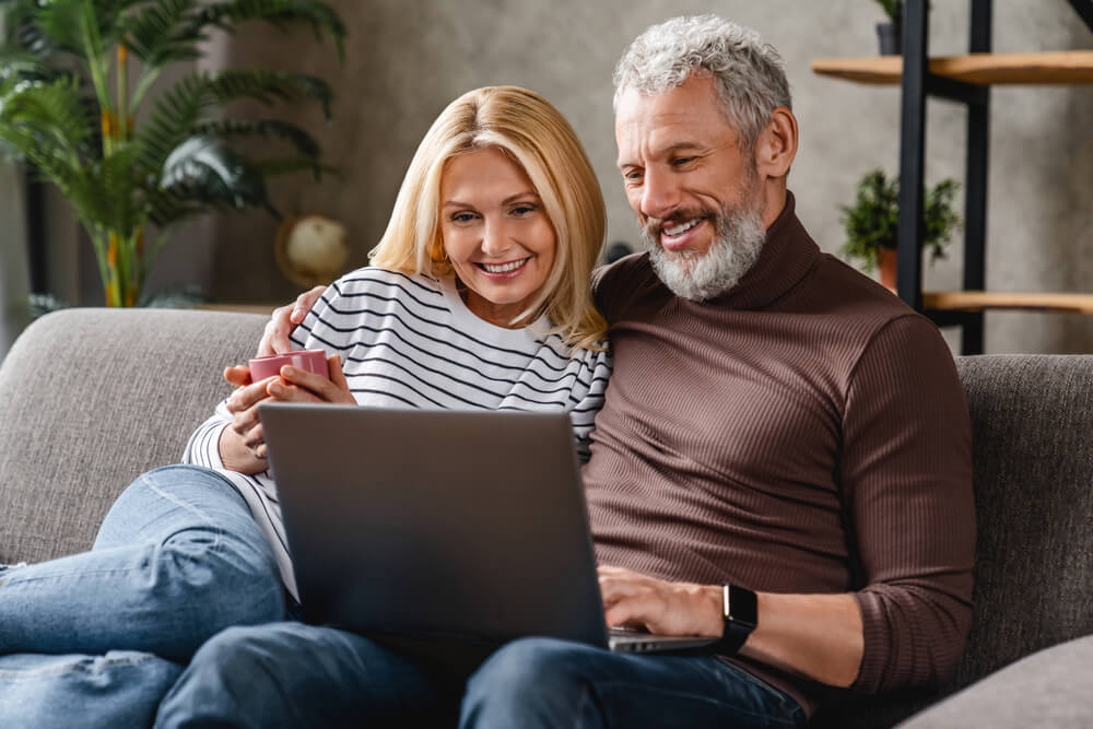 Beautiful middle aged couple using laptop and smiling while resting on couch at home