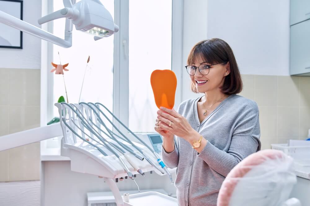 Happy woman dentist patient with mirror in hands looking at her teeth