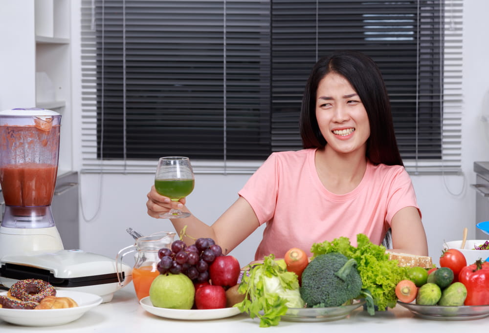 unhappy young woman drinking vegetable juice in kitchen