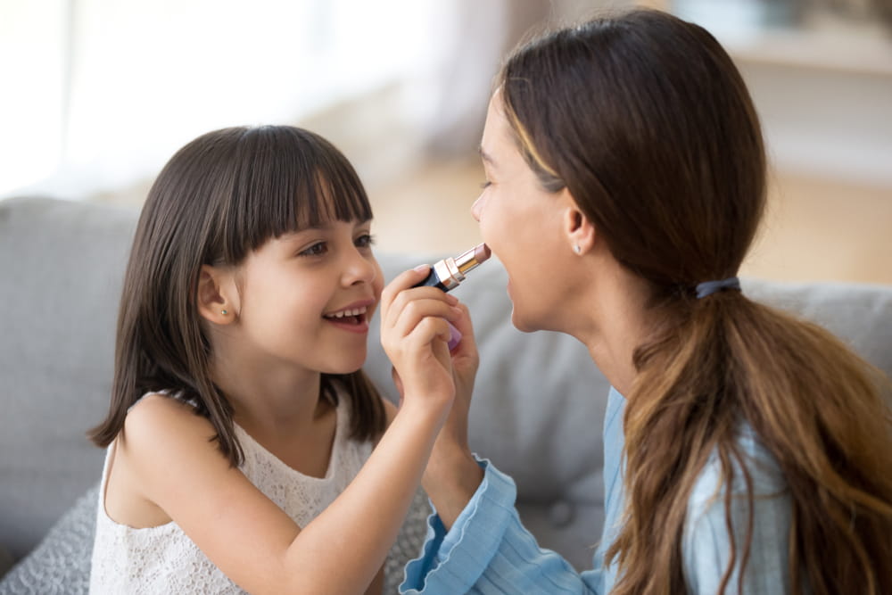 Happy young mom and cute preschooler daughter have fun together at home paint lips with lipstick,