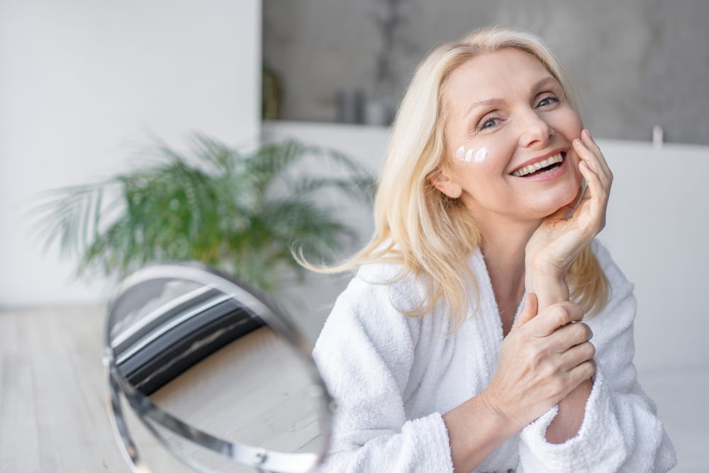 Happy, reflection and mature person with cosmetics application for glowing face in home