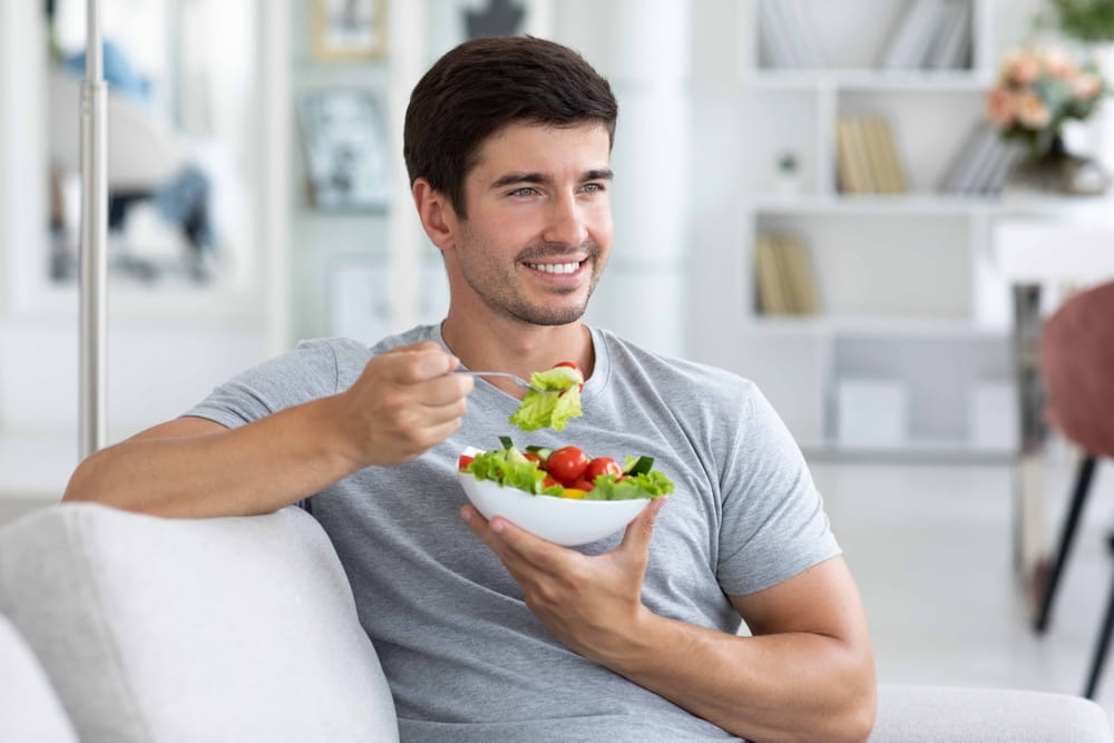Happy man sitting on sofa at home while eating green vegetable salad. Healthy eating, restaurant dish delivery.