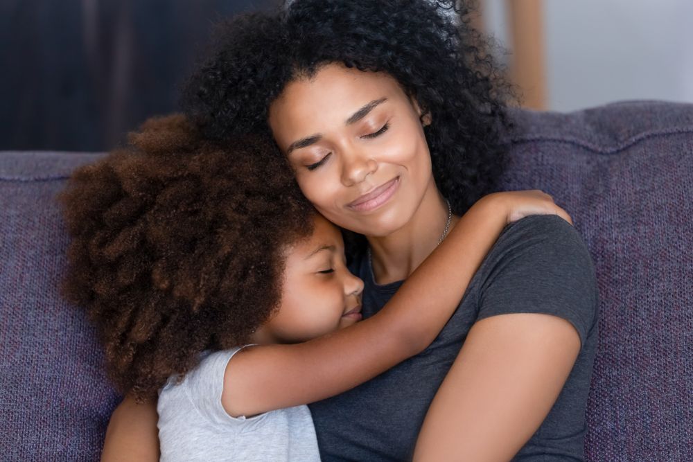 Close up biracial family portrait loving mother and little daughter sitting on couch at home hugging with closed eyes.