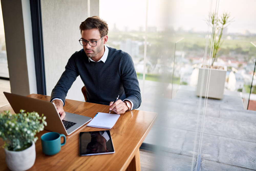 Young businessman sitting at a table at home working on a laptop and writing down ideas in a notebook