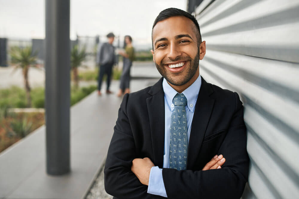 Young smiling business university student standing against college campus wall with arms crossed