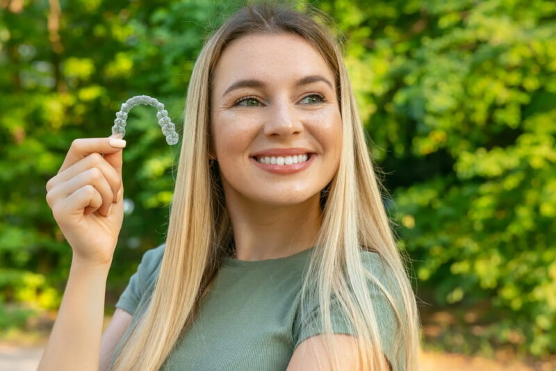 Сlose up (isolated) young beautiful woman smiling with hand holding dental aligner retainer (invisible) on green background