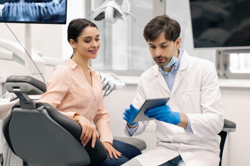 Dentist showing woman patient dental information on digital tablet while sitting in clinic, discussing treatment plan