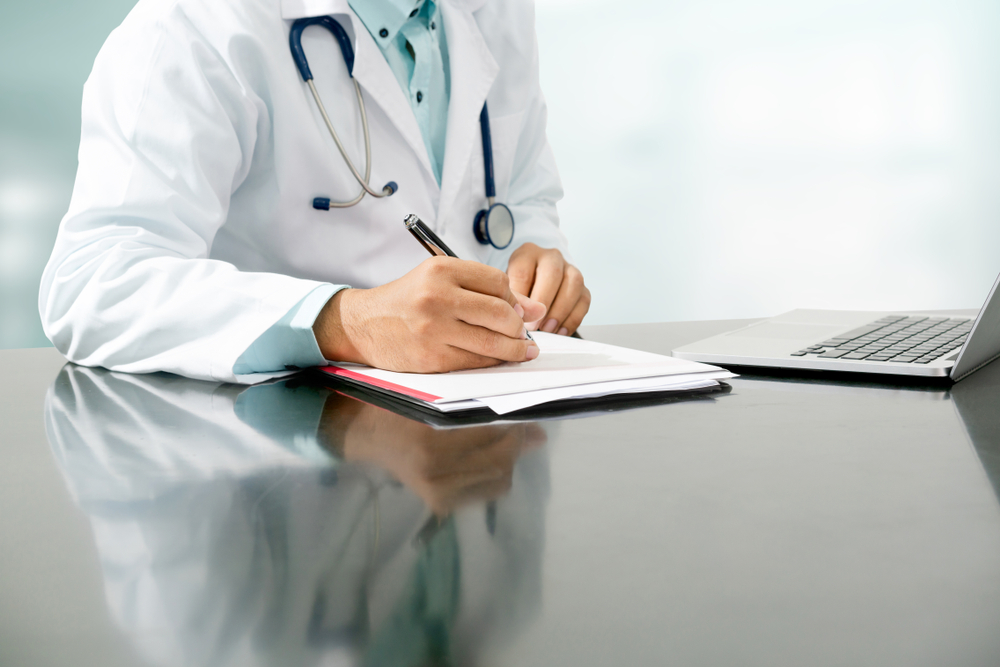 Male,Doctor,Sitting,At,Table,And,Writing,On,A,Document