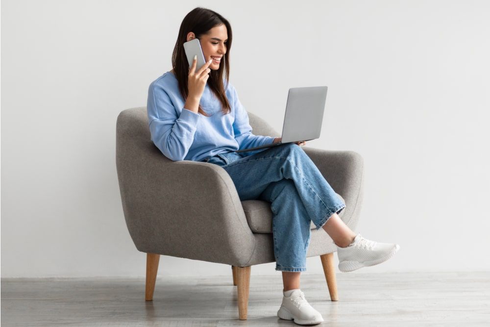 Lady using laptop and speaking on smartphone while sitting in armchair
