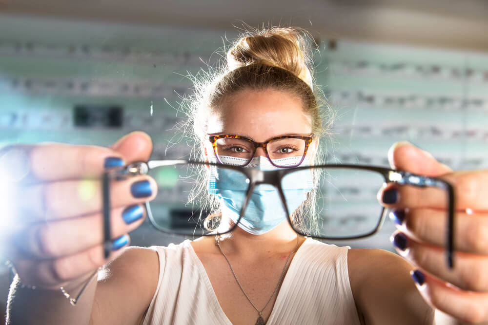 Beautiful young Optical Assistant wearing a face mask showing a pair of glasses