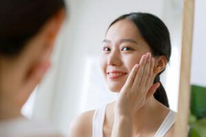 Young Asian woman touching healthy facial beauty skin look at mirror