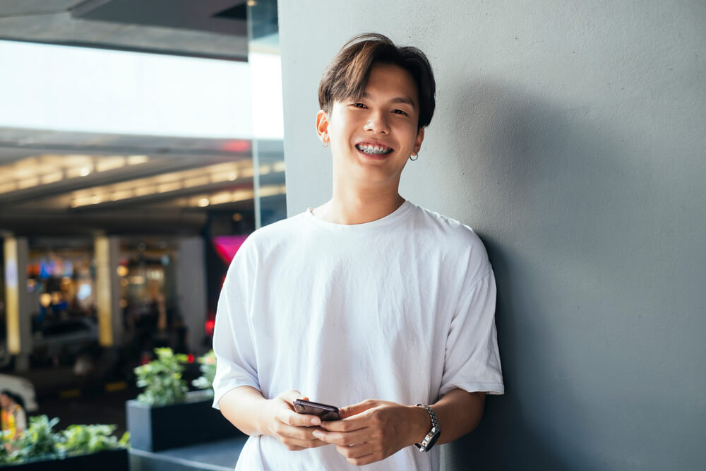 Teenager guy in white t-shirt leaning againt the building's pole using his mobile phone and smile.