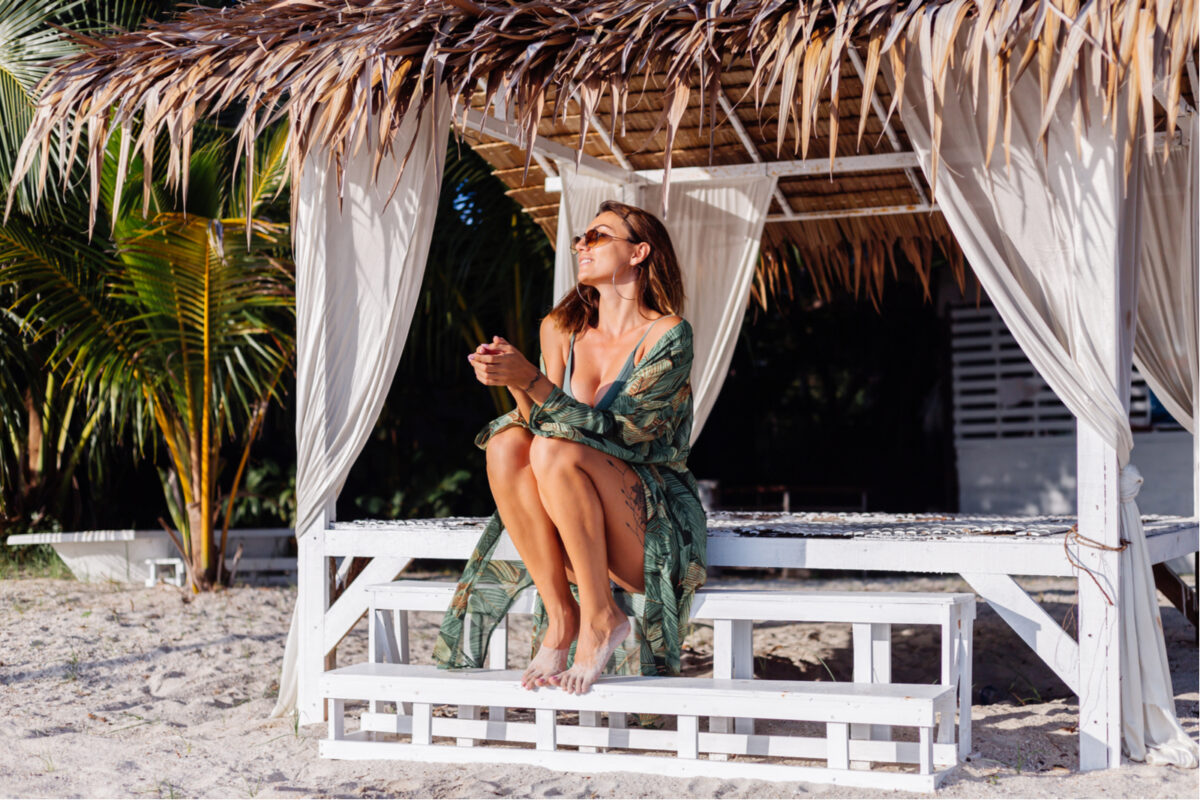 woman in denim shorts and green trendy crop top, tropical print cape, on exotic beach at sunset.