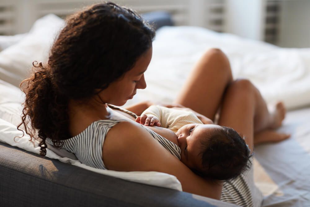 Peaceful loving young African mother sitting on bed and leaning on headboard while feeding baby