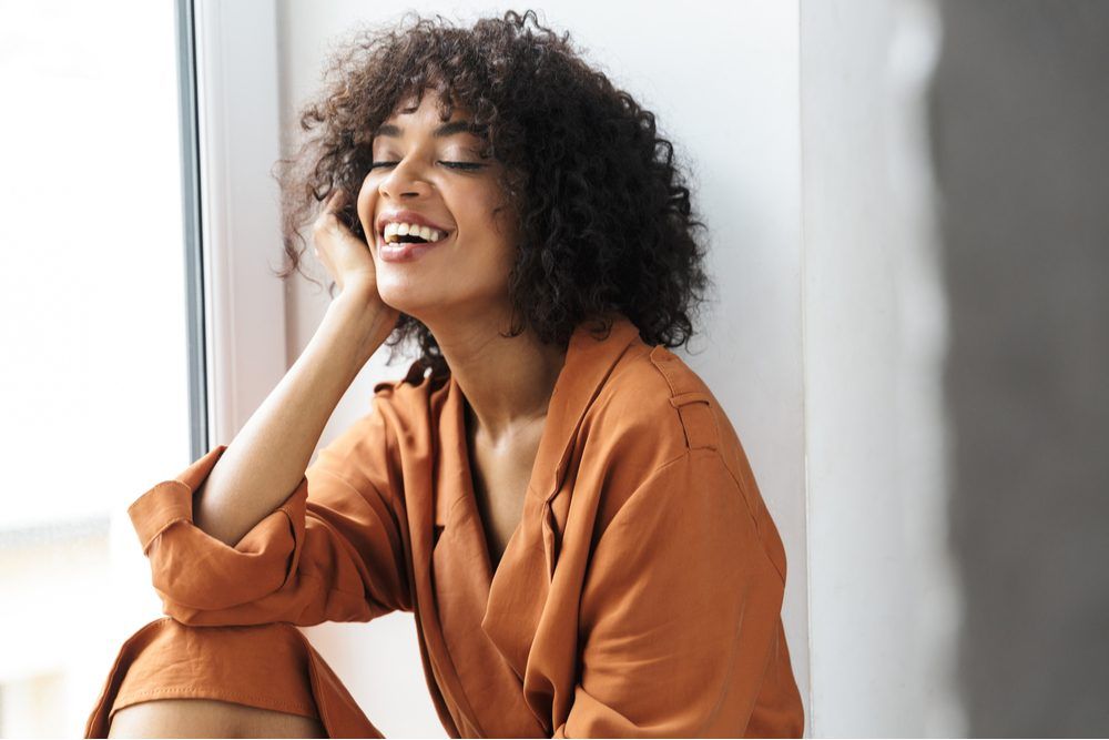 Cheerful pretty african woman having rest with closed eyes while sitting near the window