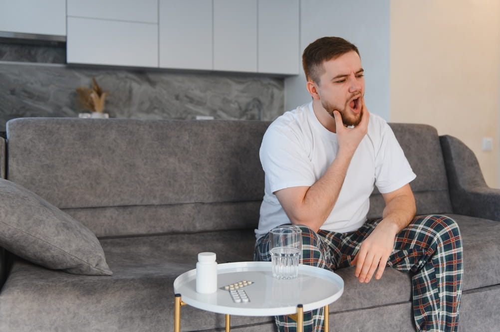 Young man experiencing severe tooth pain at home, sitting on a sofa and holding his jaw, with medication nearby