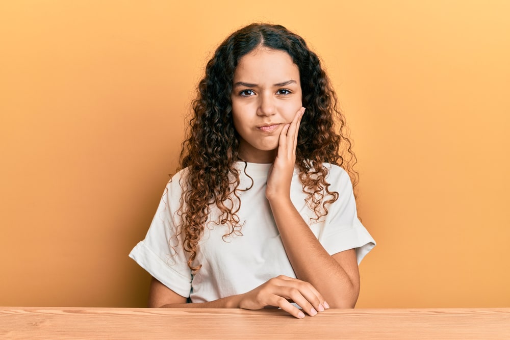 Teenager girl wearing casual clothes sitting on the table touching mouth with hand with painful expression