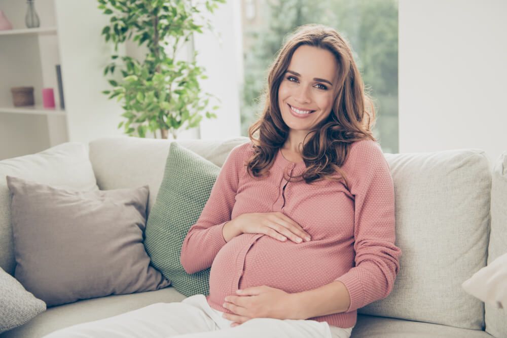 beautiful cheerful joyful pregnant curly-haired mom in casual wear sitting on divan