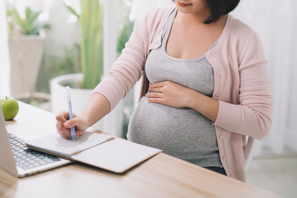 Pregnant woman writing notes and using a laptop while working