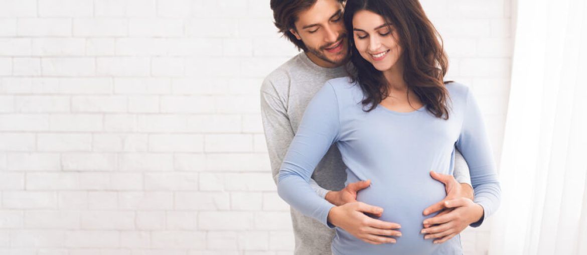 Happy couple expecting baby and hugging near window at home