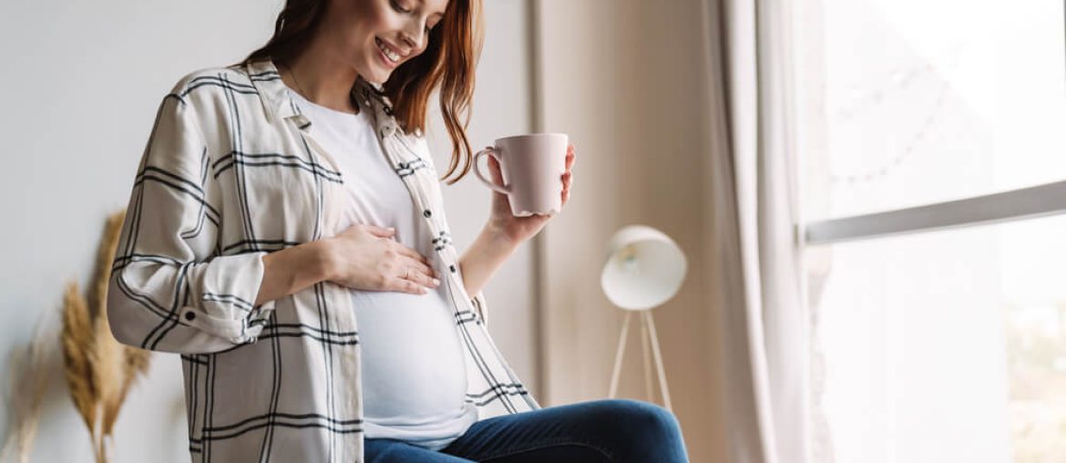 Beautiful happy pregnant woman smiling and drinking coffee while sitting on couch at home