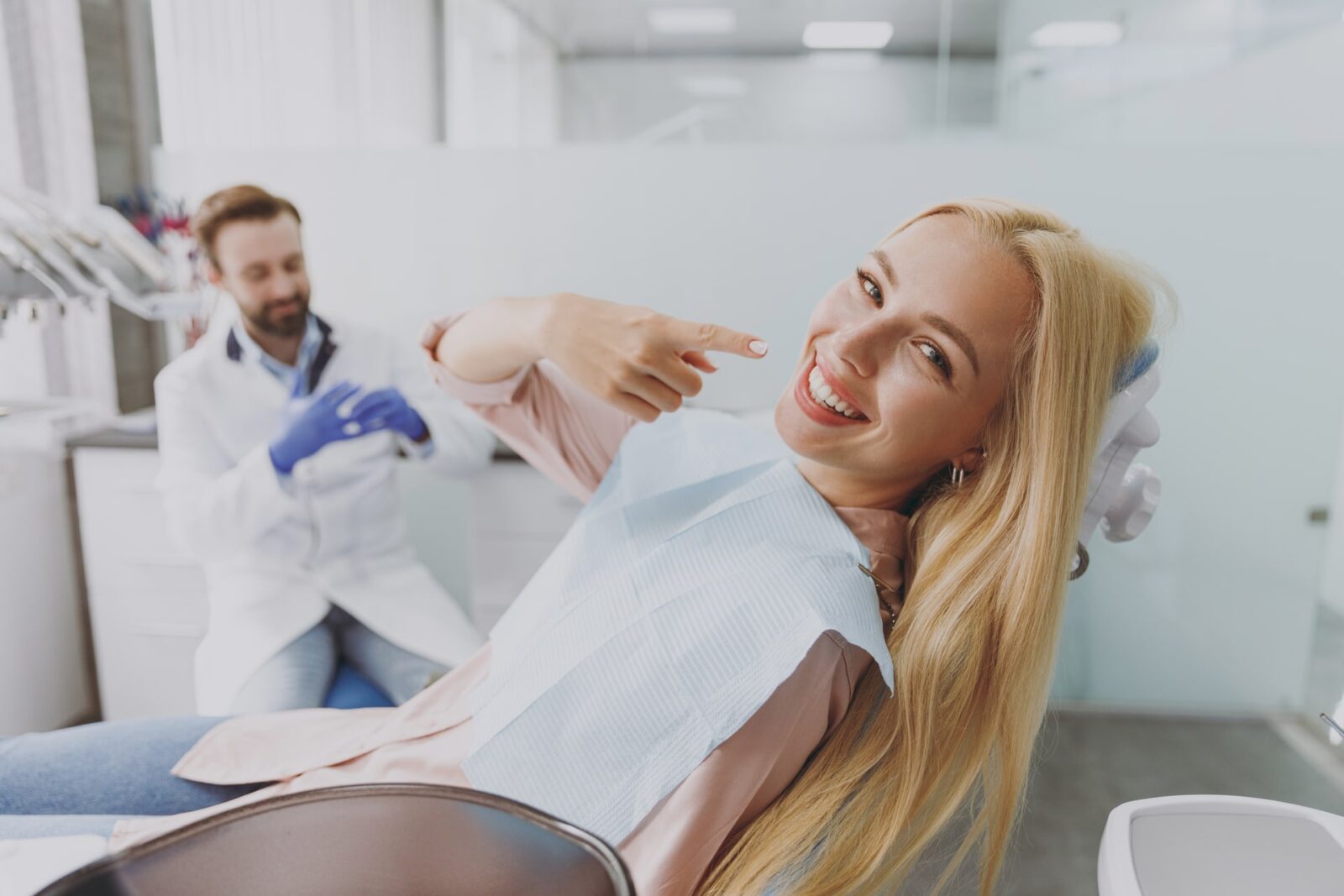 Young smiling happy woman with dental napkin point finger on mouth sit at dentist office chair