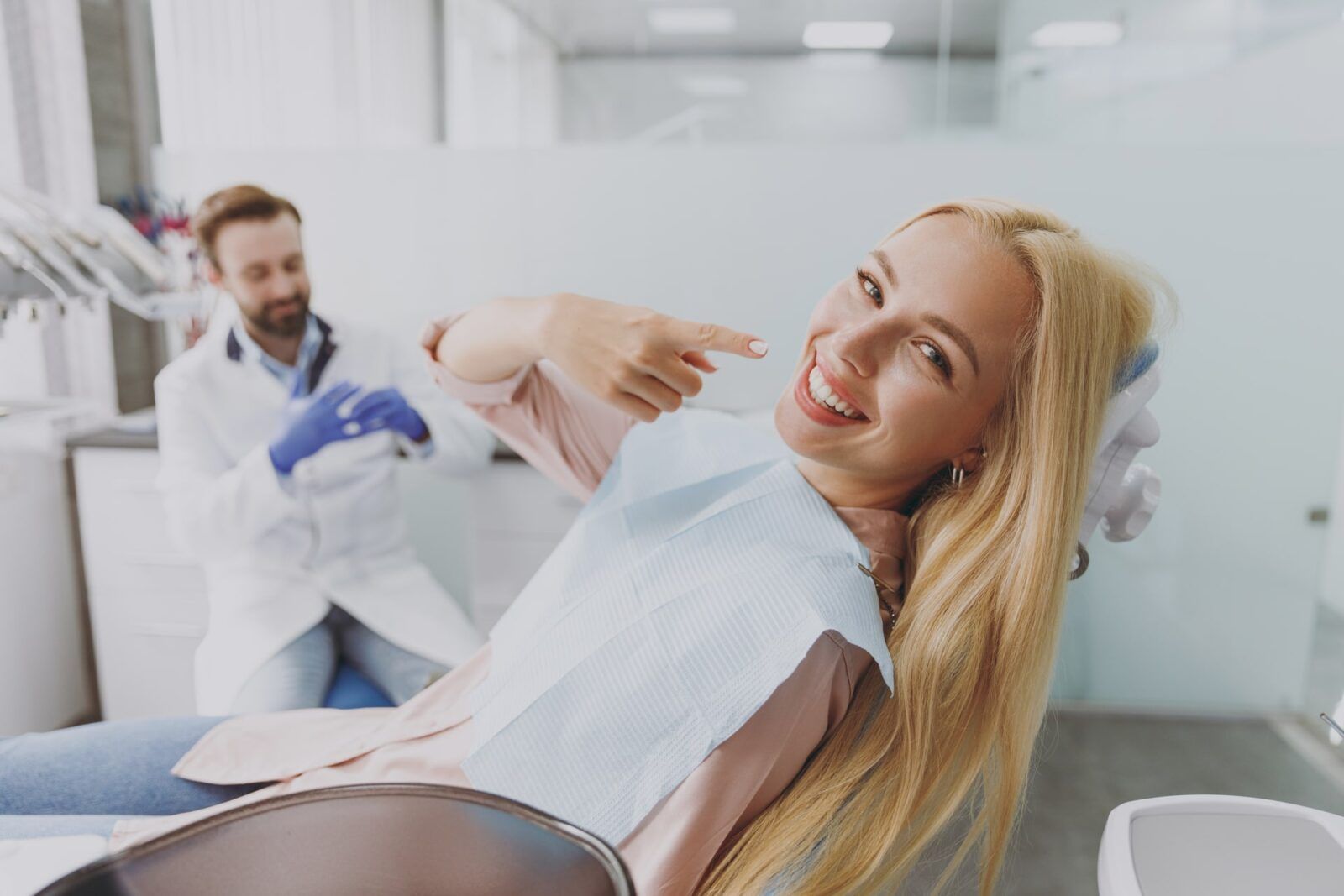 Young smiling happy woman with dental napkin point finger on mouth sit at dentist office chair