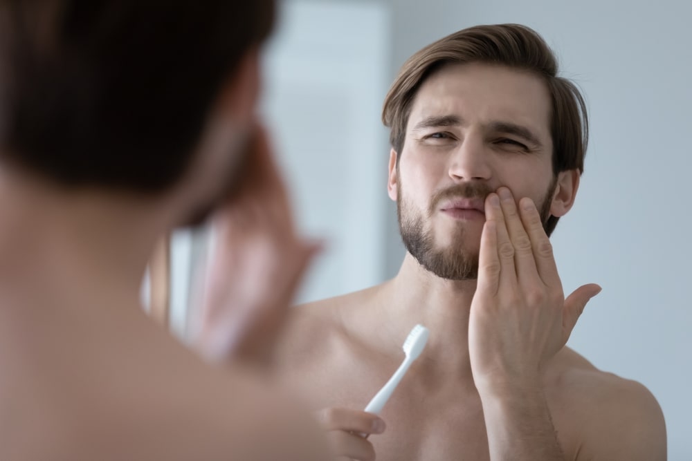 man brushing his teeth and holding his mouth because he is in pain