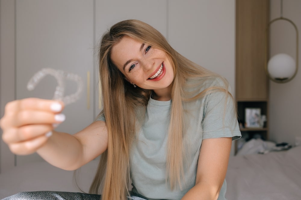 A cheerful woman proudly displays clear aligners, highlighting modern dental care and orthodontic solutions, in a cozy home interior.