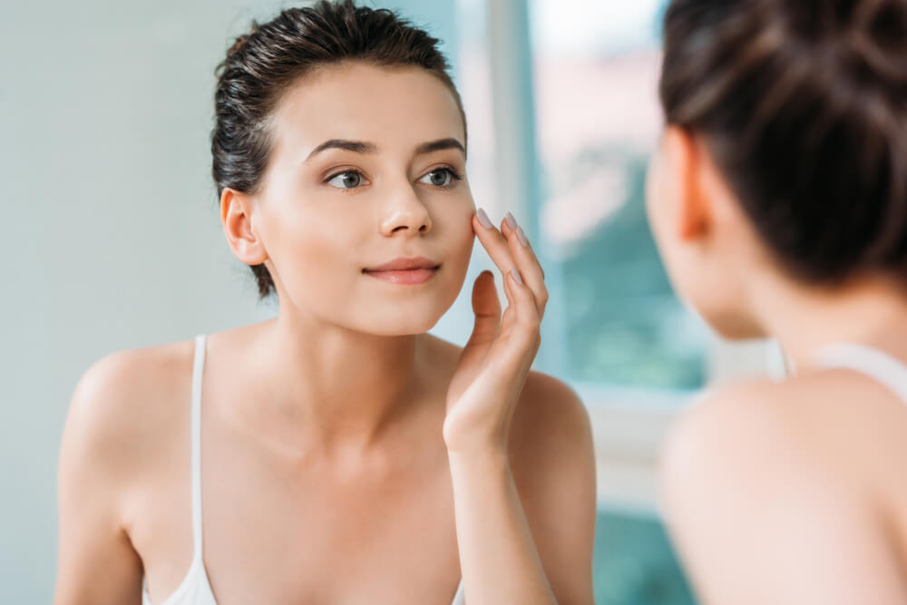 beautiful smiling young woman touching face and looking at mirror in bathroom