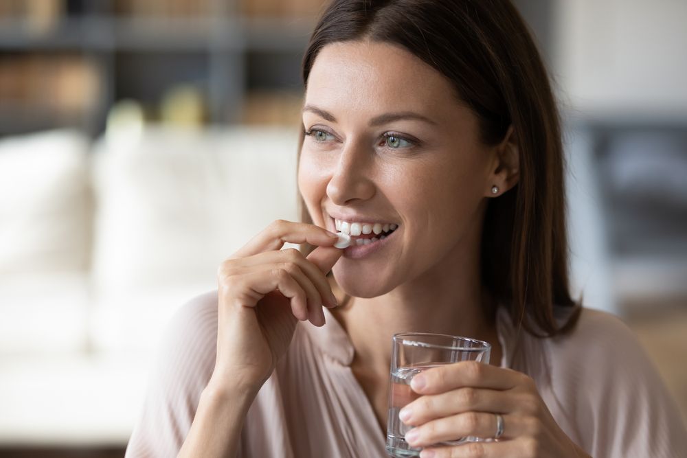 smiling woman taking white round pill, holding water glass in hand