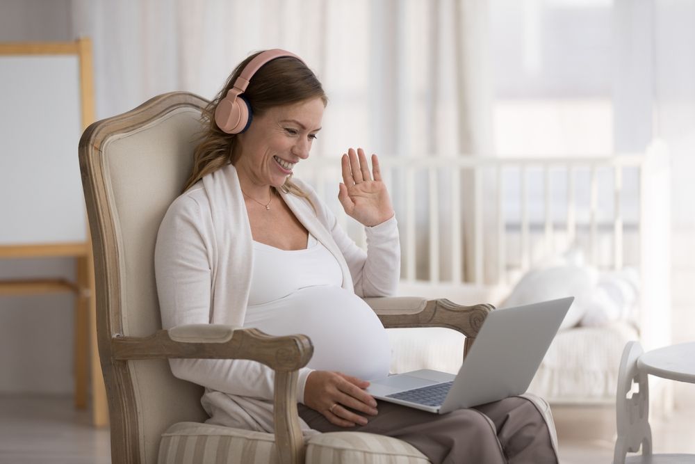 Pregnant woman sit in armchair with laptop wear headphones wave hand start video call with obstetrician-gynecologist.