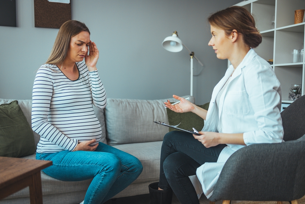 Young pregnant woman talking with doctor and having headache. Woman during a routine check up with her doctor at home