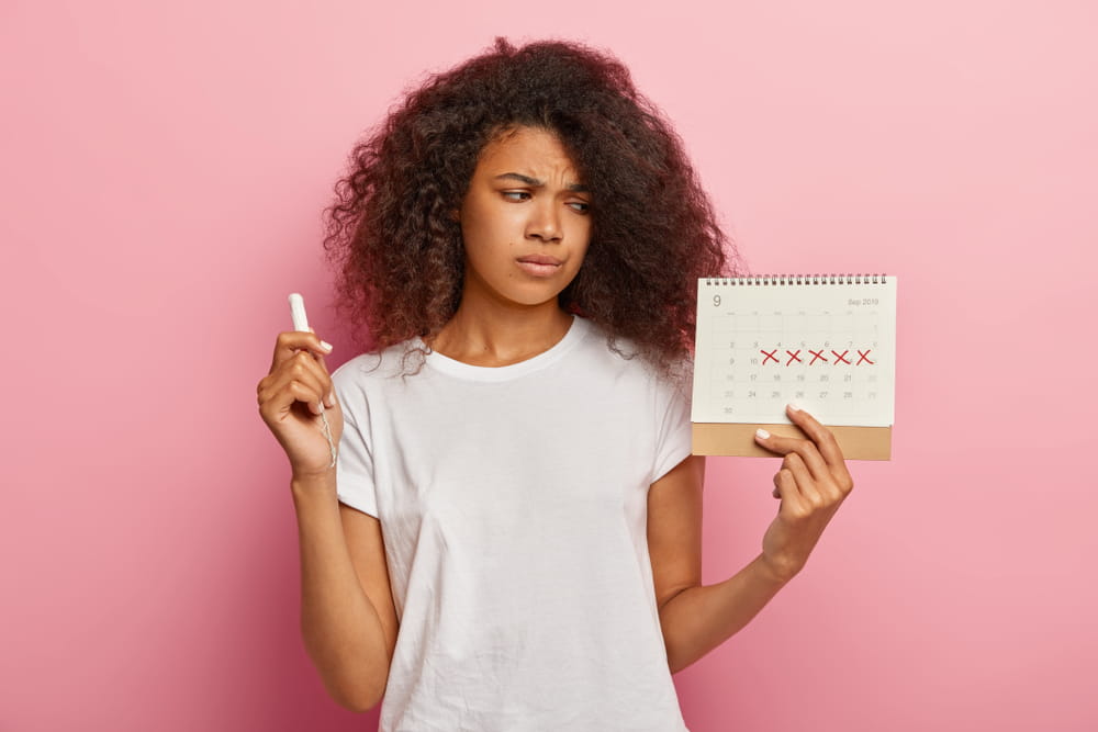 Displeased stressful dark skinned woman looks at periods calendar with marked red crosses, has painful menstruation, holds tampon for good protection