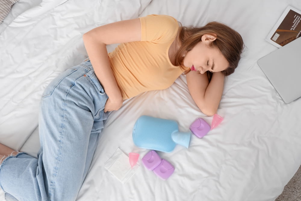 Young woman suffering from menstrual cramps with feminine hygiene items on bed, top view