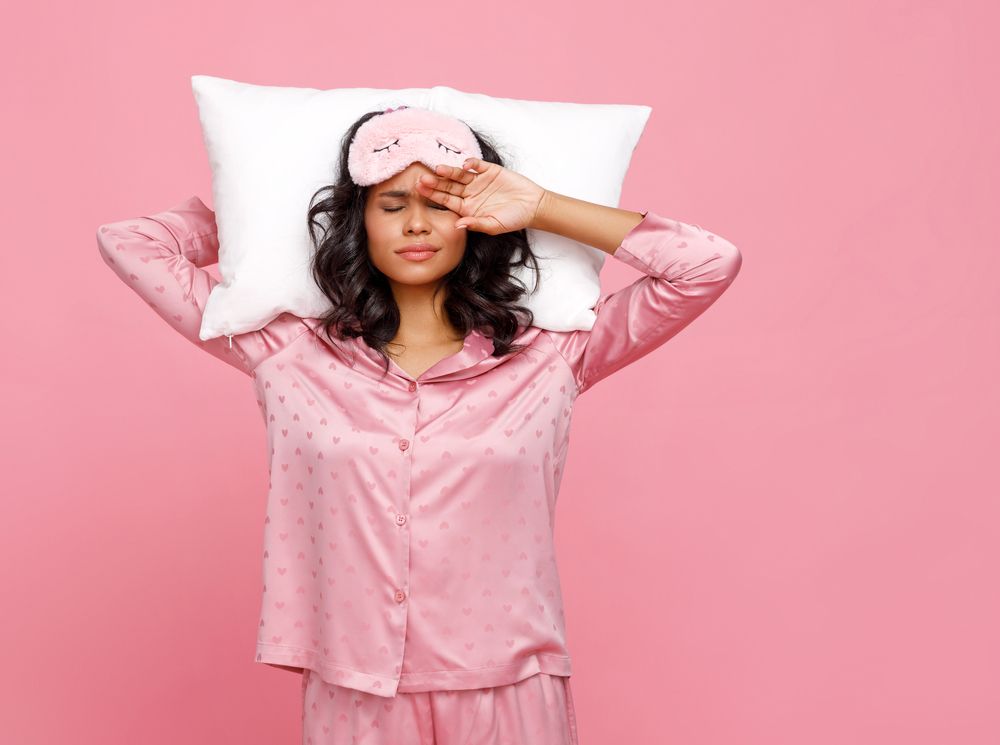 woman in sleepwear and mask holding pillow behind head and rubbing face