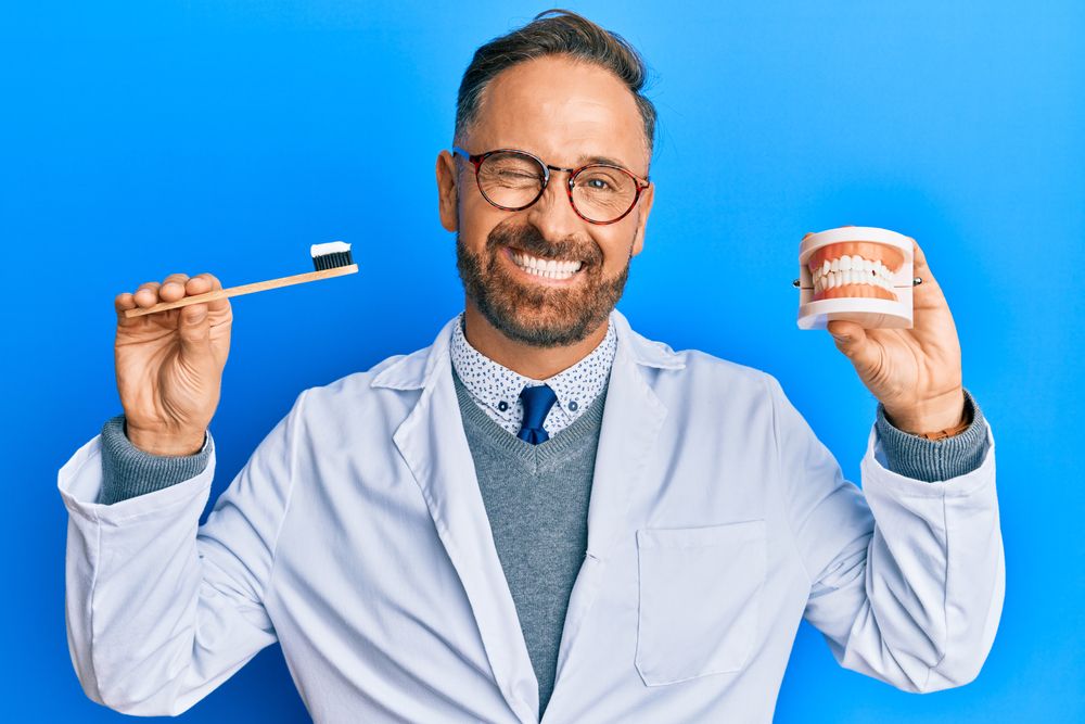 Handsome middle age dentist man holding denture and toothbrush winking looking
