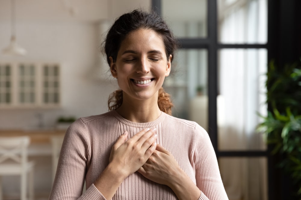 Happy peaceful millennial hispanic lady stand alone with closed eyes keep hands close to heart