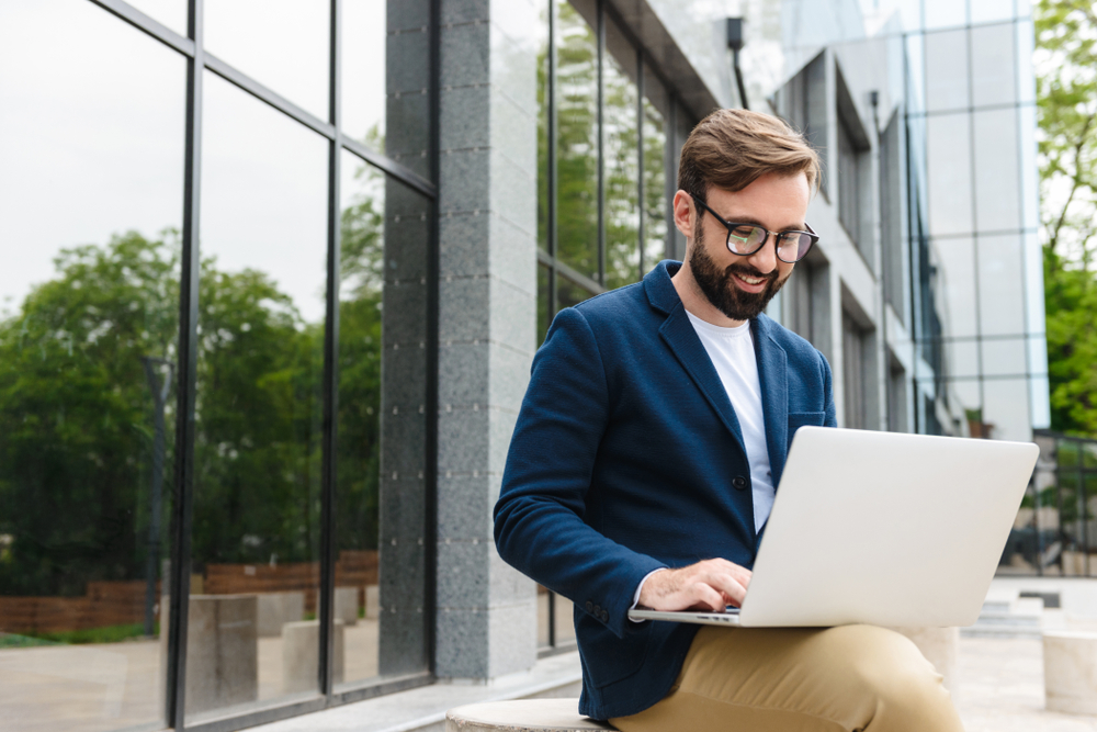 Attractive smiling young bearded man wearing jacket working on laptop