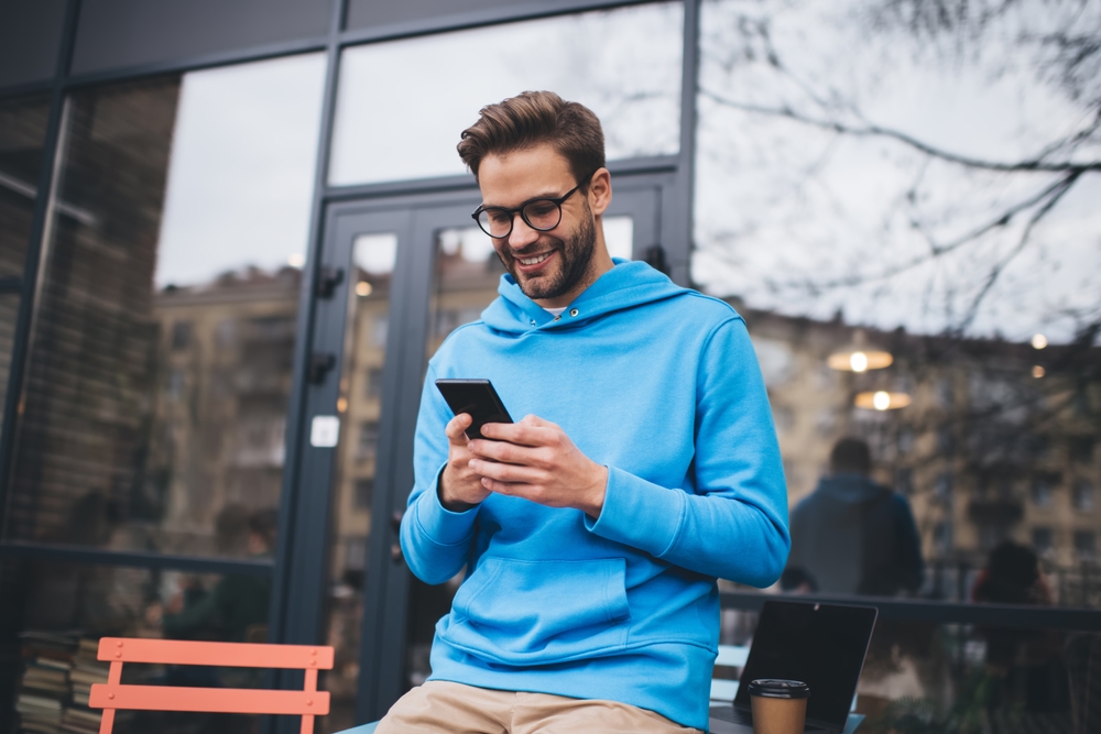 Happy hipster guy in classic spectacles for provide vision correction using cellphone