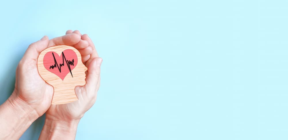 Hands gently cradling a wooden cutout of a human head with a heart and lifeline graphic inside, illustrating the comprehensive care and maintenance needed for neurological rehabilitation after an injury.
