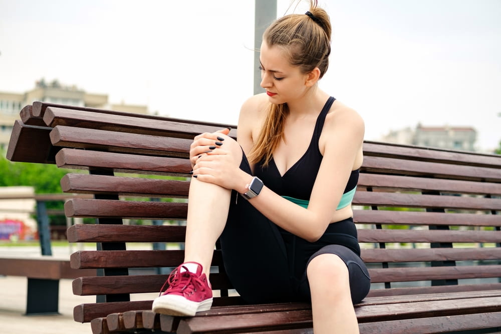 Young brunette woman wearing sportive clothes on city park, outdoors cramps in leg and feel pain, touching her knee ankle