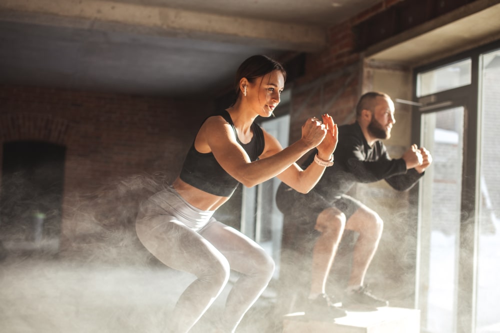 couple doing squats on boxes in a crossfit style gym, well lit by bright sunlight.
