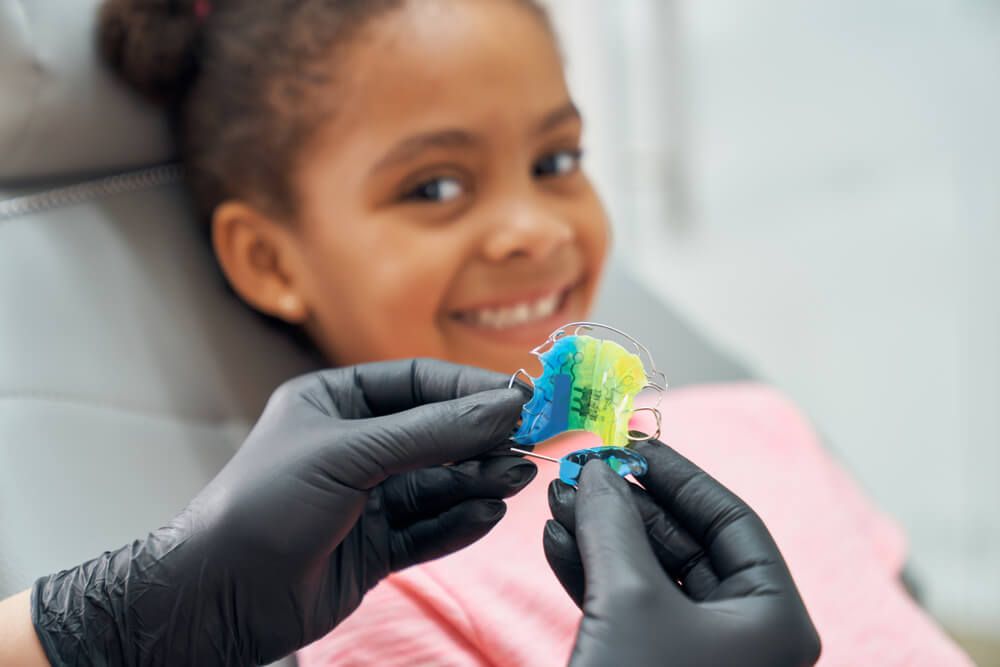 Hands of dentist in black rubber gloves holding colorful braces. Pretty girl sitting in dental chair