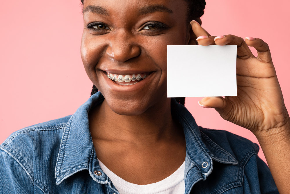 Orthodontics. African American Girl In Dental Braces Showing Orthodontist's Visiting Card Posing Over Pink Background. Mockup