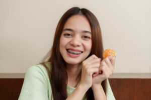 Asian teenage girl with long brown hair holding peeled orange, showing braces, smiling brightly.