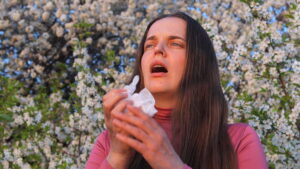 A woman stands amidst blooming trees, visibly affected by pollen allergies while holding a tissue in her hands during spring.