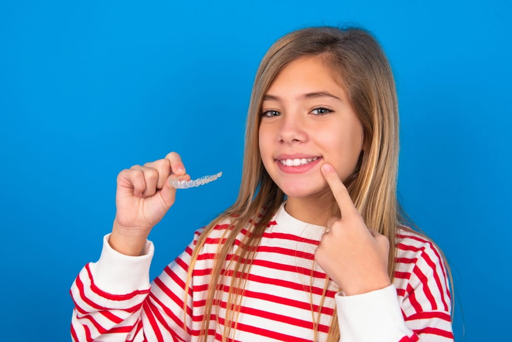 beautiful caucasian teen girl wearing striped shirt over blue studio background holding an invisible aligner